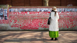 a woman stand in front of a wall decorated with red hearts and photos of people who died during the Covid-19 pandemic